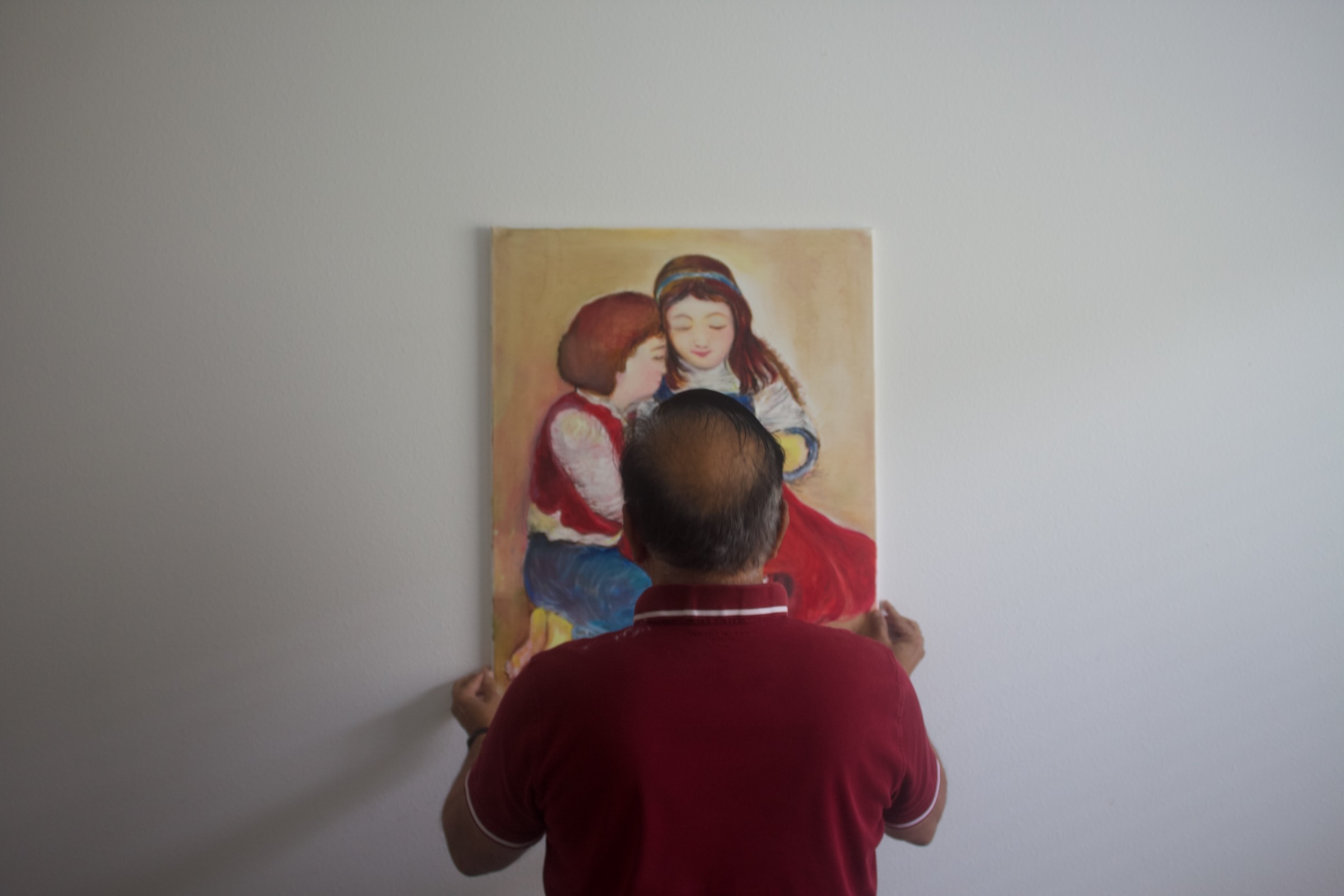 Person in a red shirt seen from behind while hanging a painting of two seated children.